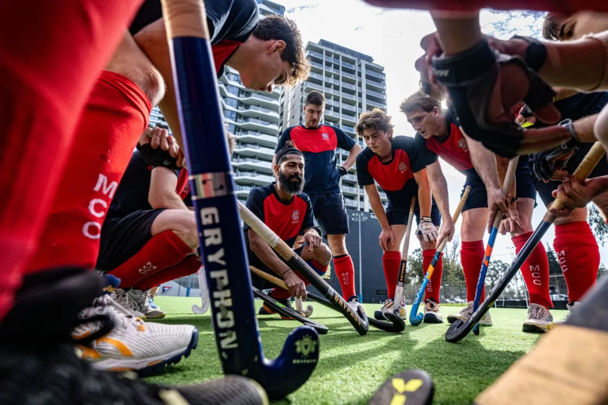 A field hockey team in red and black uniforms huddles together on an outdoor turf, holding their sticks and listening to a coach.
