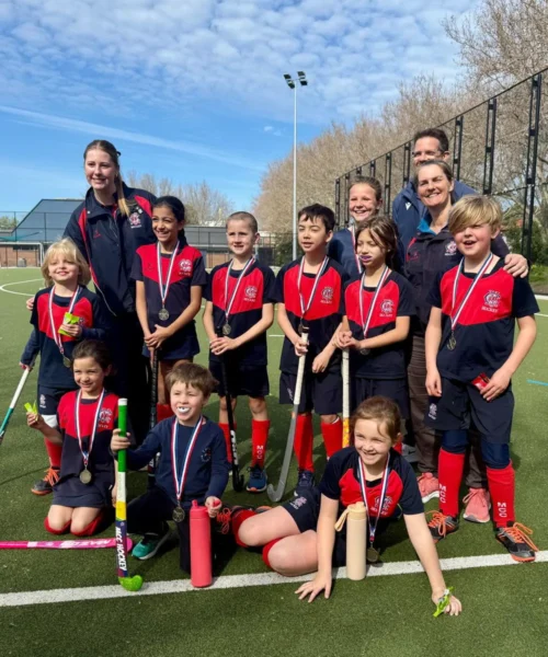 A youth field hockey team poses on an outdoor field with coaches, all wearing medals and uniforms, smiling for a group photo