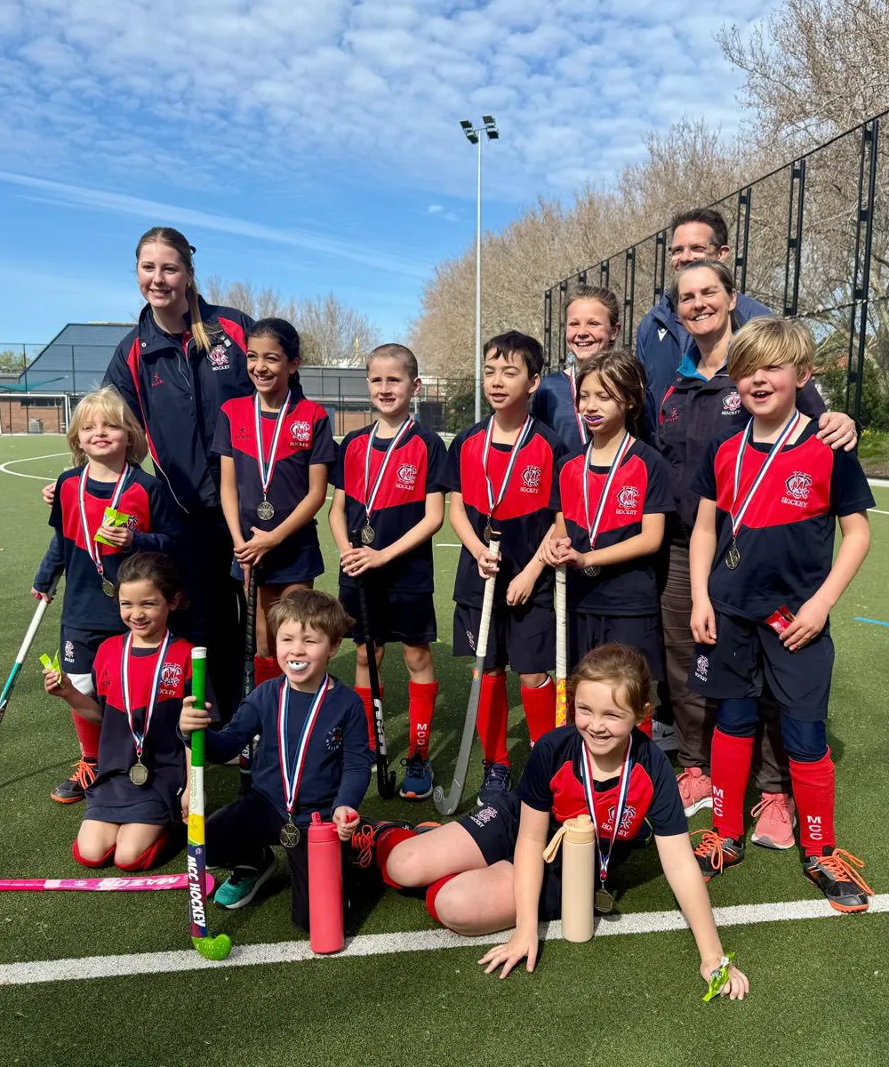 A youth field hockey team poses on an outdoor field with coaches, all wearing medals and uniforms, smiling for a group photo